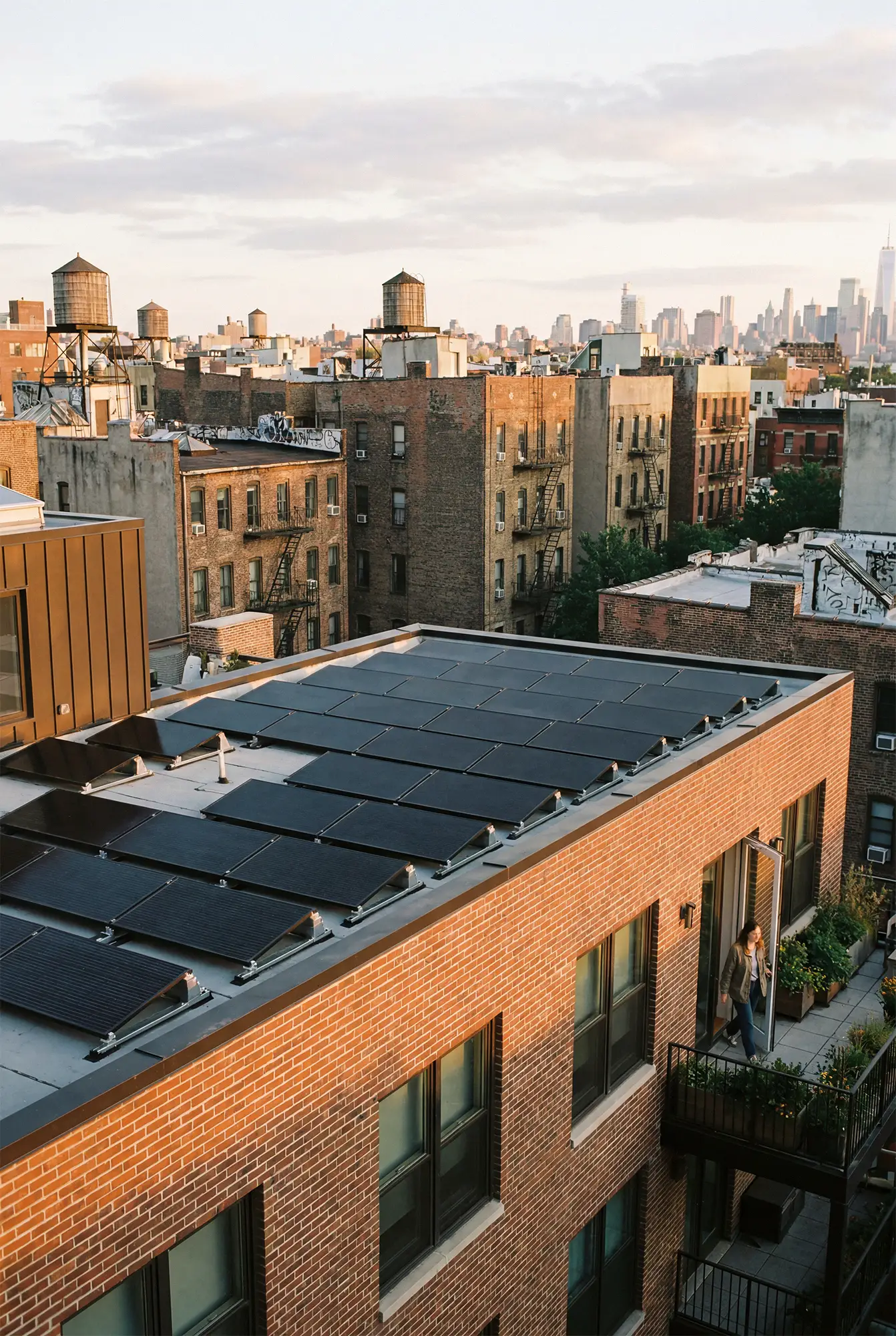 Rooftop solar panels on a brick apartment building in a New York city neighborhood, with more buildings and water towers in the background at sunset.