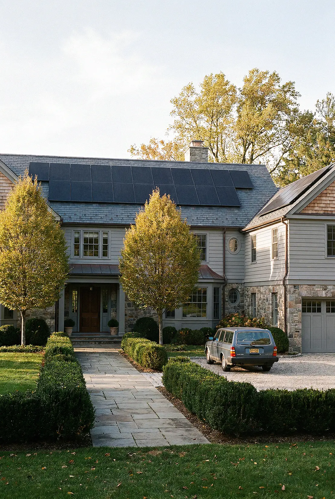 A two-story house with solar panels on a New York roof, flanked by two trees in front, with a blue station wagon parked on a gravel driveway.