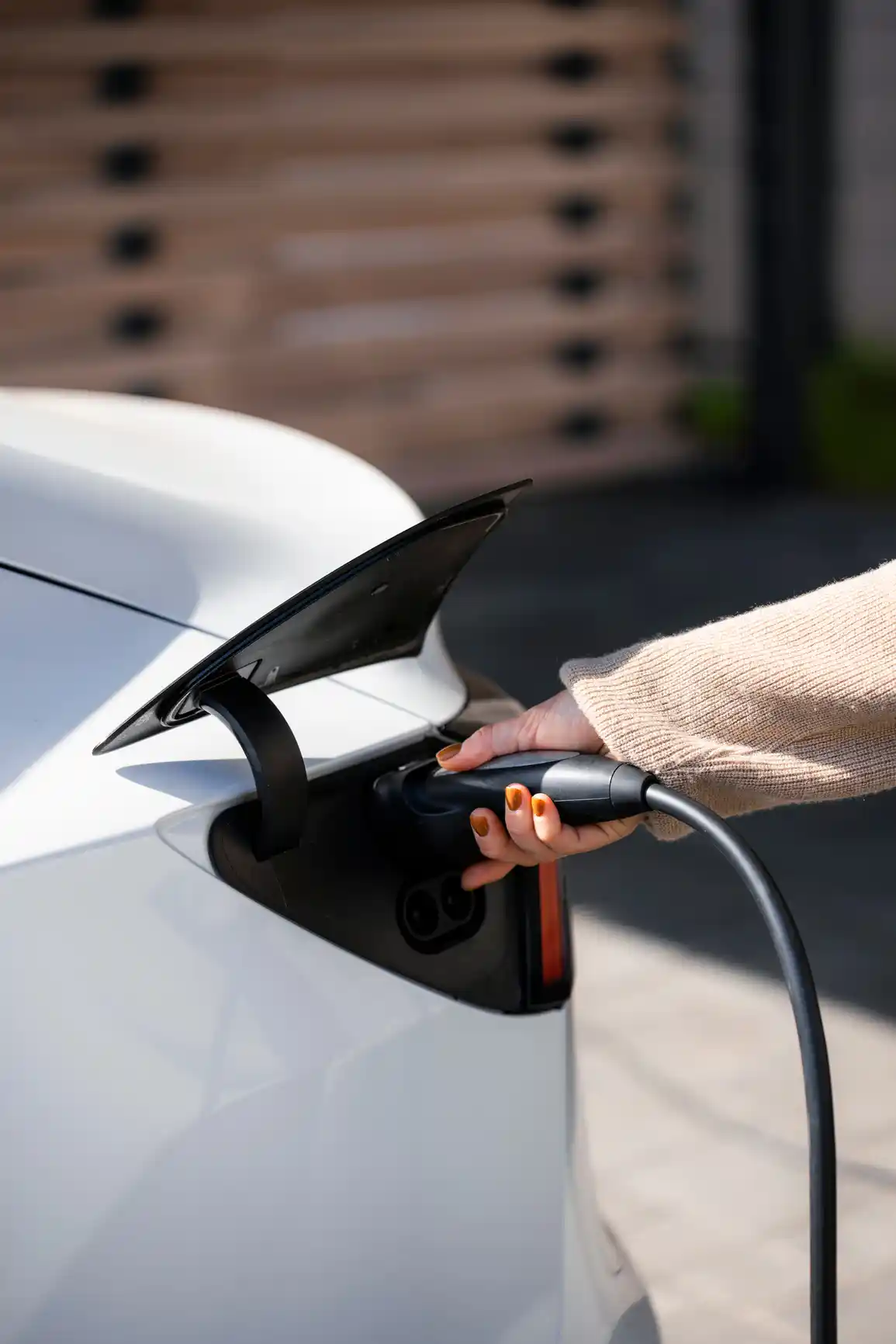 A person connects an electronic vehicle charger to a white electric car. The charging port is open, revealing their hand holding the cable. Dressed in a beige sweater with orange nail polish, they stand against a blurred background.