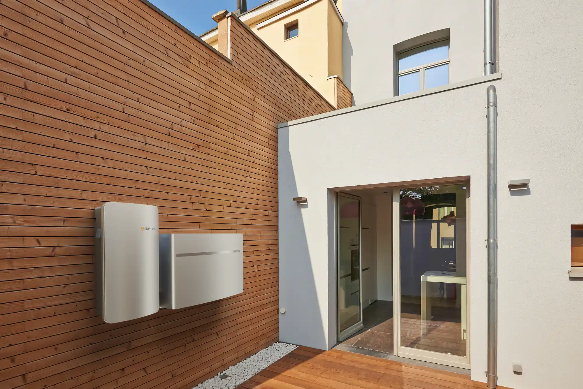 A white building with glass doors opens onto a wooden deck. Solar panels enhance the roofline, seamlessly integrating green energy solutions.