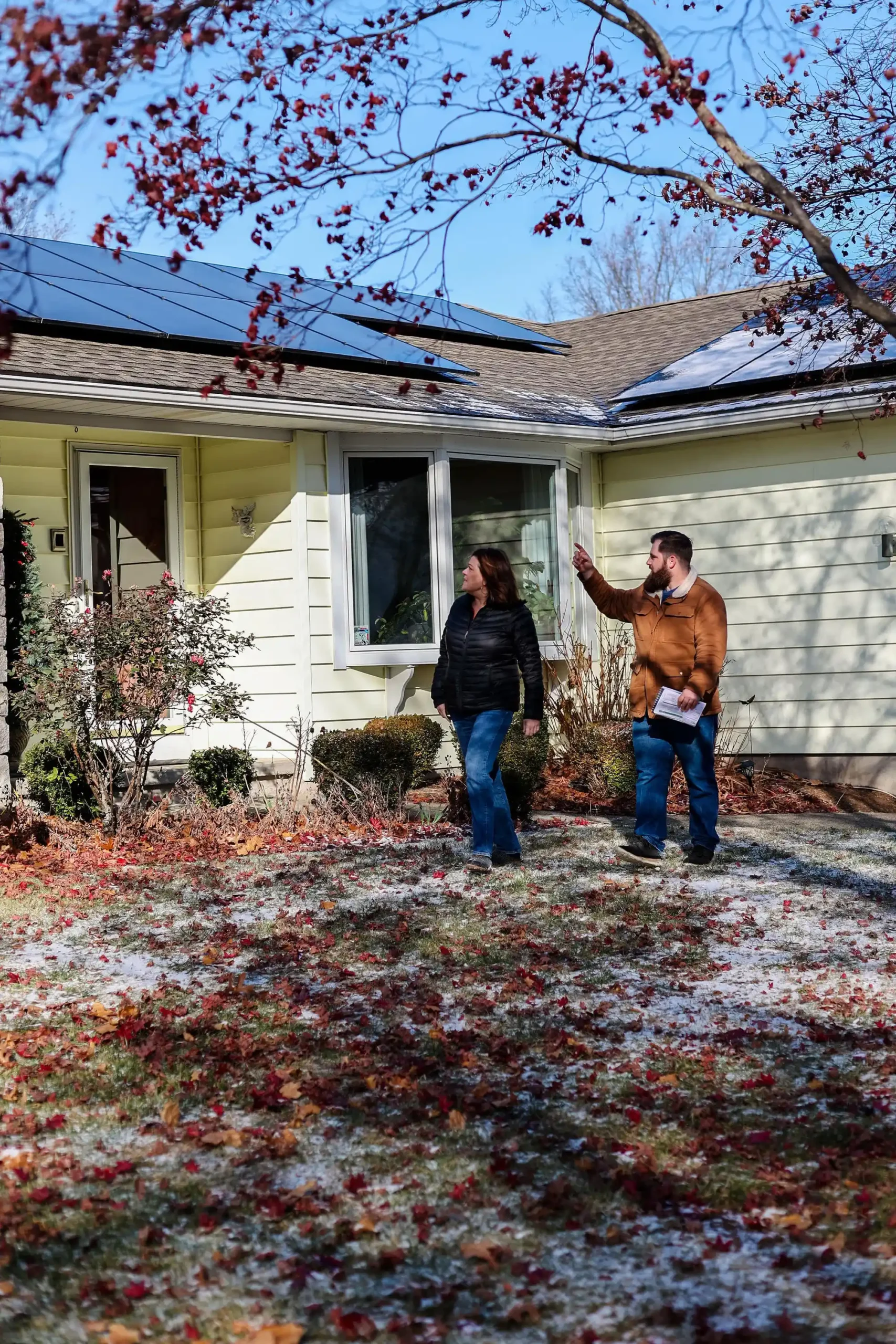 A man and a woman stand in front of a house with solar panels and smart electrical panels on the roof. Theyre both wearing jackets and jeans, surrounded by sparse patches of snow and fallen leaves. The woman looks towards the man, who is pointing at the roof.