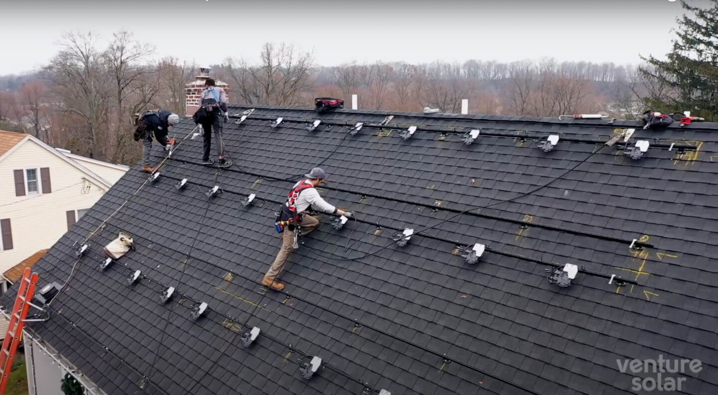 Workers wearing safety gear expertly install solar panels on a dark shingled, steeply pitched roof equipped with brackets for the installation. Trees and a clear sky frame the scene, with Venture Solar visible in the corner. A seamless battery backup installation enhances efficiency.