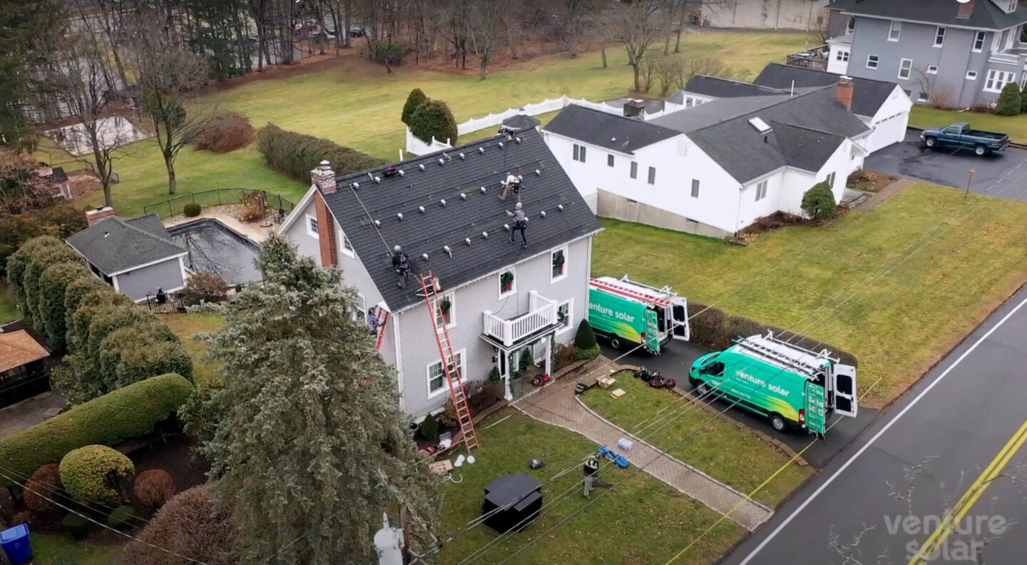 Venture Home vehicles parked outside a large home while workers install solar panels on the roof.