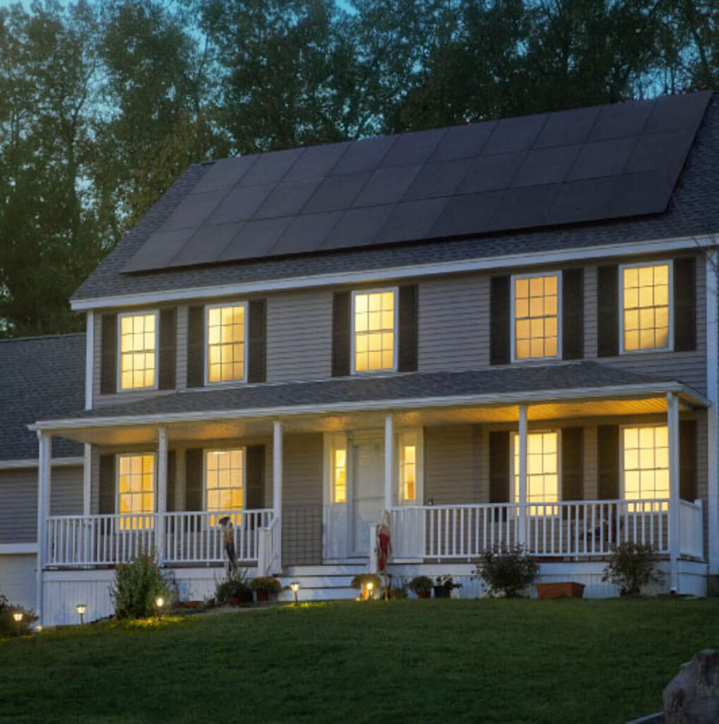 A two-story house with solar panels on the roof and a reliable battery backup is illuminated from within, set against a backdrop of trees. The home features a spacious front porch, multiple lit windows, and a lush grassy lawn in the foreground.