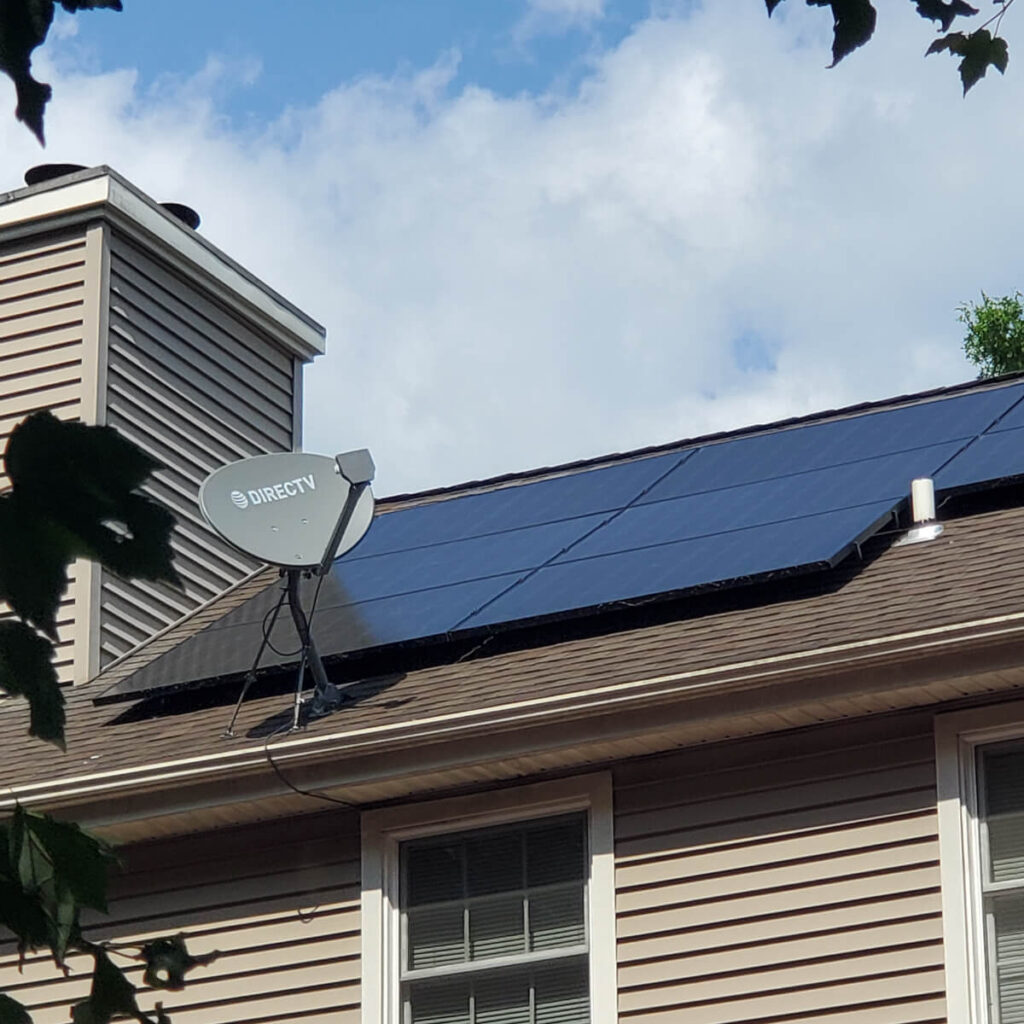 Solar panel installation and a satellite dish grace the roof of a house with beige siding and white-trimmed windows. The sky is partly cloudy, while tree leaves frame the scene, hinting at the homes readiness for energy independence.