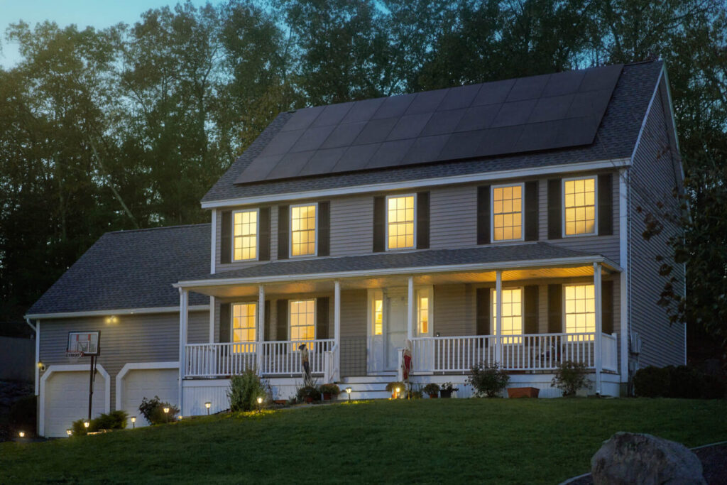 A cozy two-story house with a porch and expertly done solar panel installation on the roof is lit warmly against a twilight sky. Tall trees form the backdrop. A well-kept lawn and a basketball hoop in the driveway enhance the inviting atmosphere, now secure with battery backup installation.