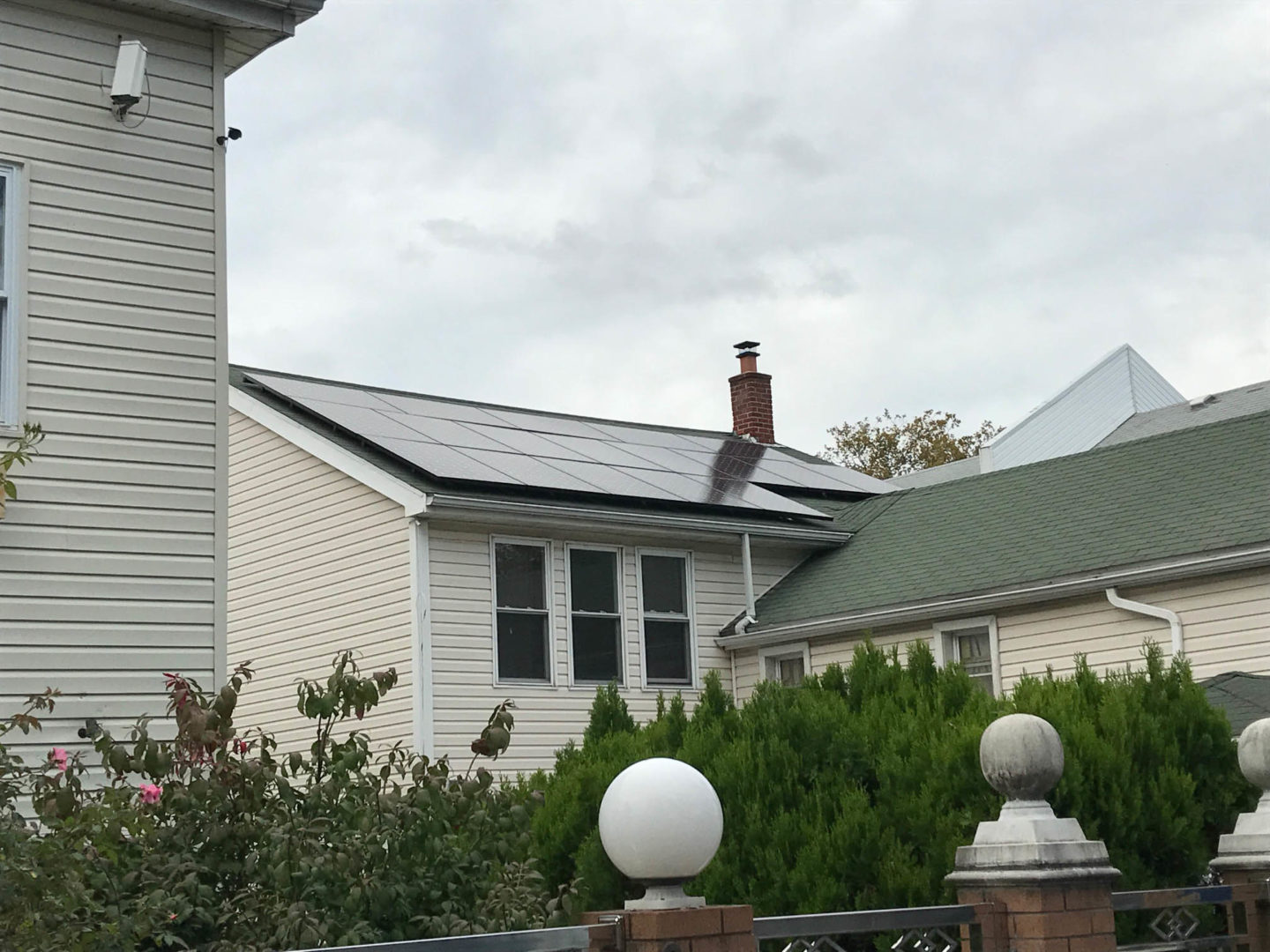A home with solar panels installed on the roof, surrounded by greenery and a white fence with decorative pillars.