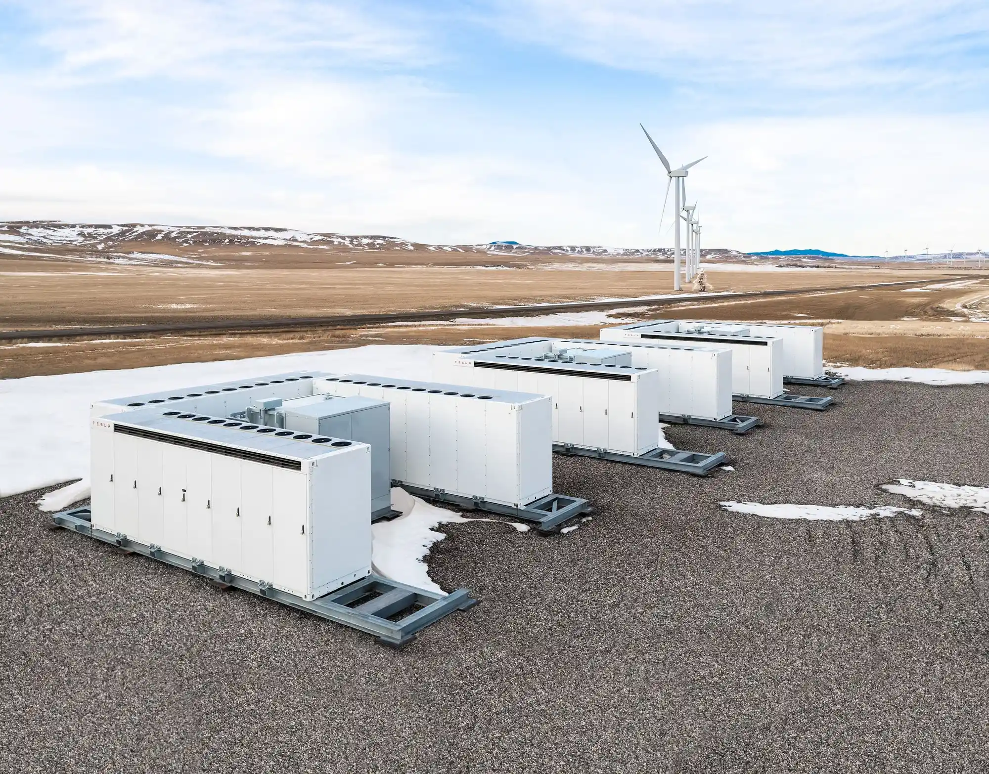 Four white industrial battery storage units sit on a gravel surface with patches of snow. In the background, a single wind turbine rises over open fields and hills under a cloudy sky, highlighting the potential for integrating commercial solar projects into this renewable energy landscape.