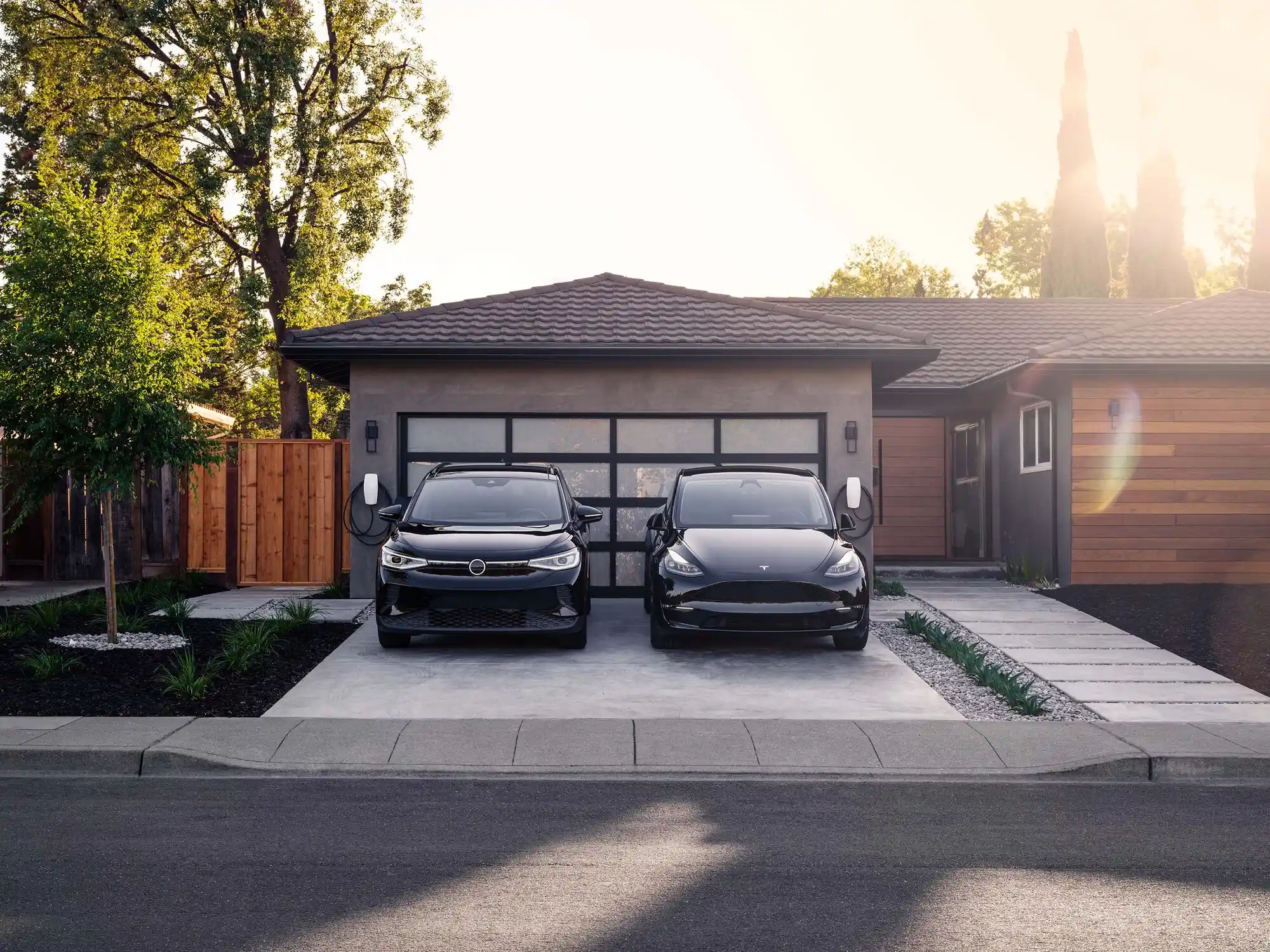 Two modern electric SUVs are parked side by side in the driveway of a contemporary house with a wooden exterior and glass garage door. With trees framing the scene, this eco-friendly home, benefiting from local solar incentives, showcases neatly arranged plants and a wooden fence.