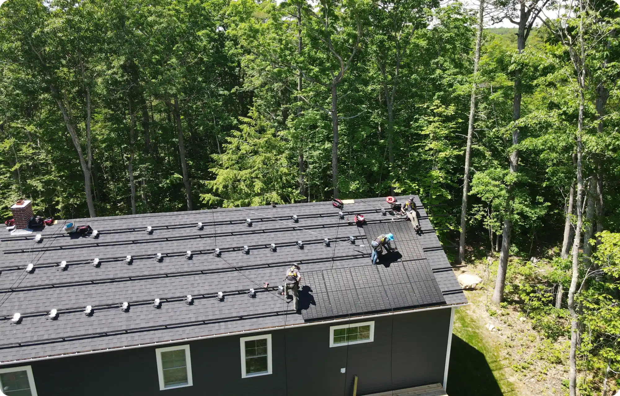 Workers are busy with a commercial solar installation on the black roof of a large building, surrounded by dense green trees. Equipment and materials are scattered around them as they work tirelessly under bright daylight in this forested area.