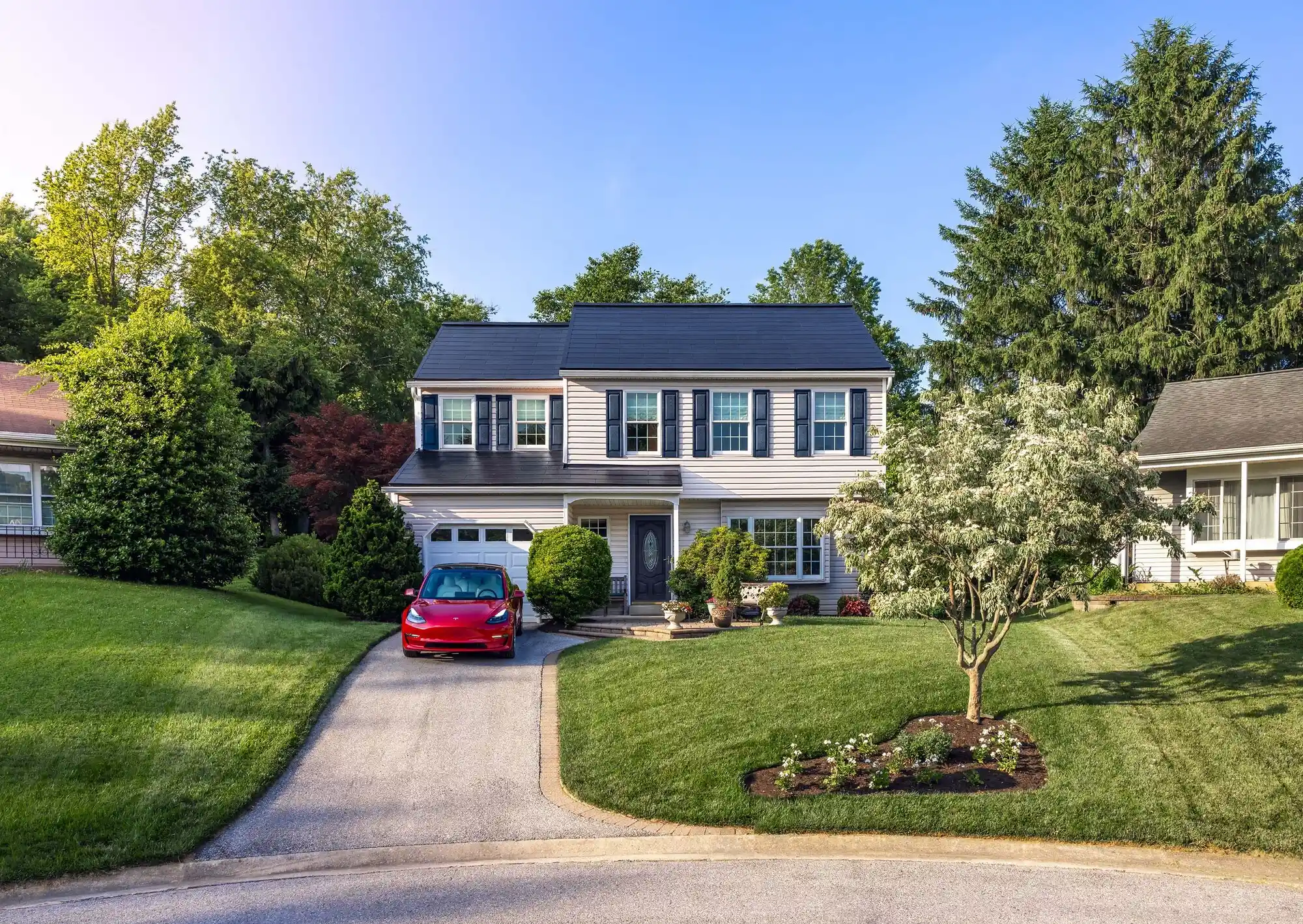 A suburban two-story house with beige siding and black shutters sits under a clear blue sky. Alongside the red car in the driveway, solar panels blend seamlessly into the roof, showcasing its commitment to clean energy among lush green lawns and manicured shrubs.