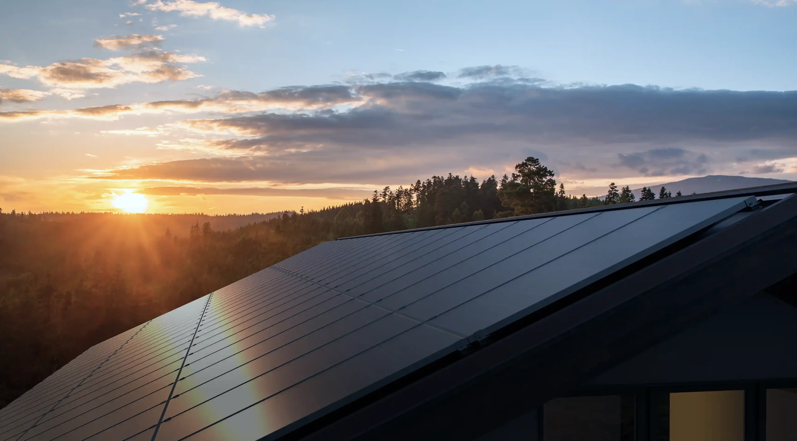 A rooftop covered with solar panels, expertly installed, is seen under a sunset sky. The sun casts a warm glow across the horizon, partially obscured by scattered clouds. Forested hills are visible in the background, perfectly setting the scene for your venture home.