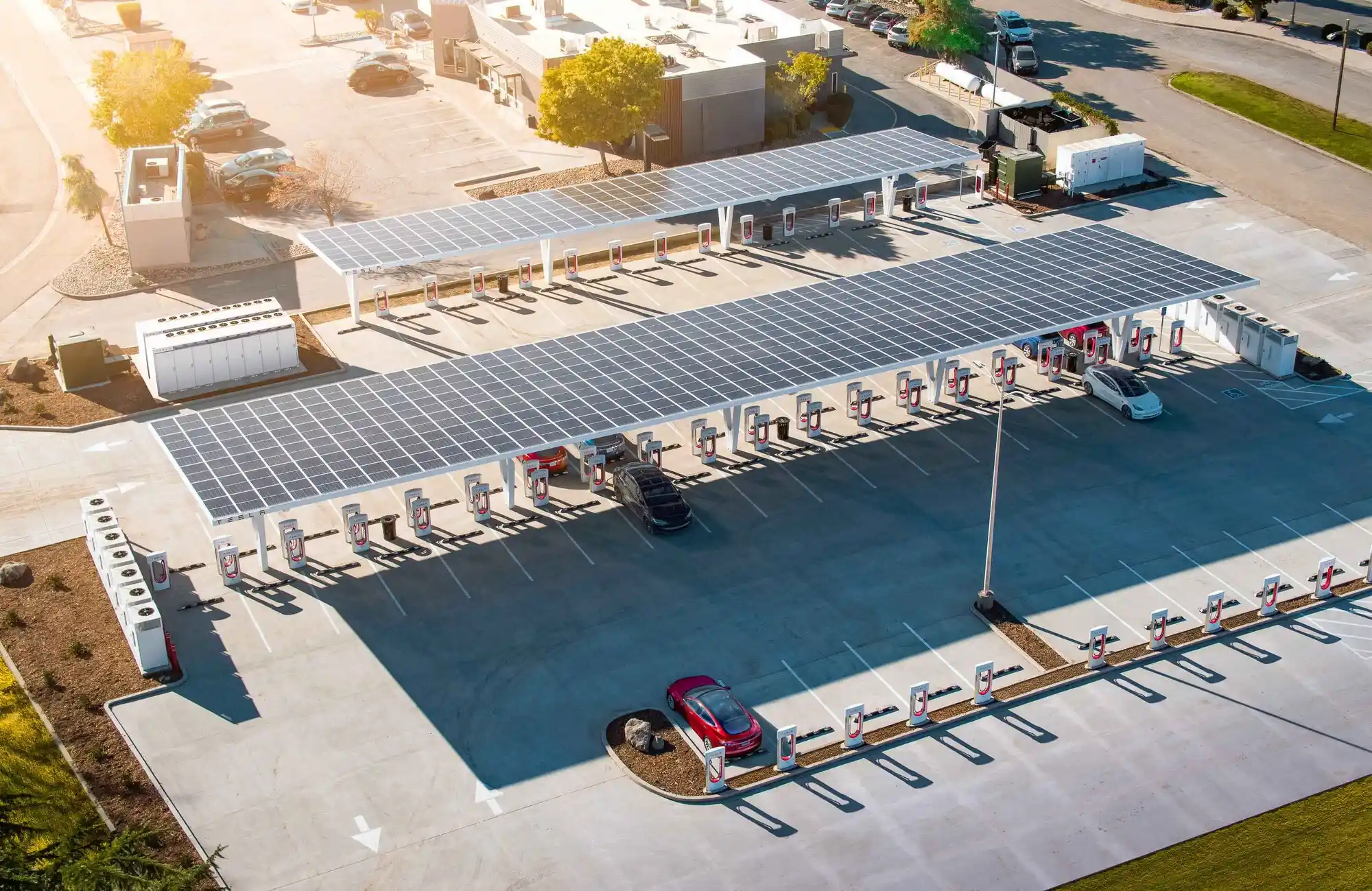 Aerial view of a commercial solar installation at an electric vehicle charging station with solar panel-covered canopies. Multiple charging stalls are visible, with two cars parked and charging amid nearby buildings, trees, and a clear sky above.