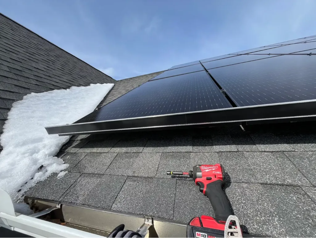 A power drill rests on a shingled roof beneath solar panels, with melting snow nearby, hinting at the need for snow guards under the clear blue sky.