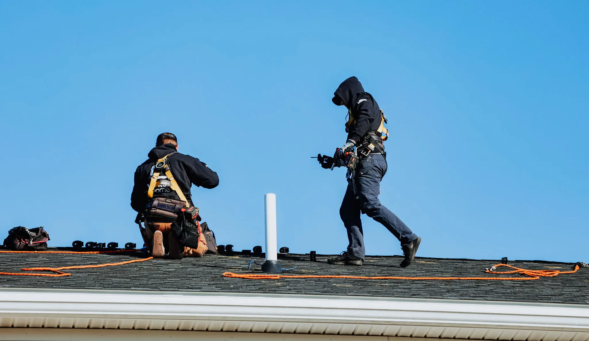 Two workers in safety gear are focused on roof installation or repair, using tools and orange safety ropes, set against a clear blue sky.