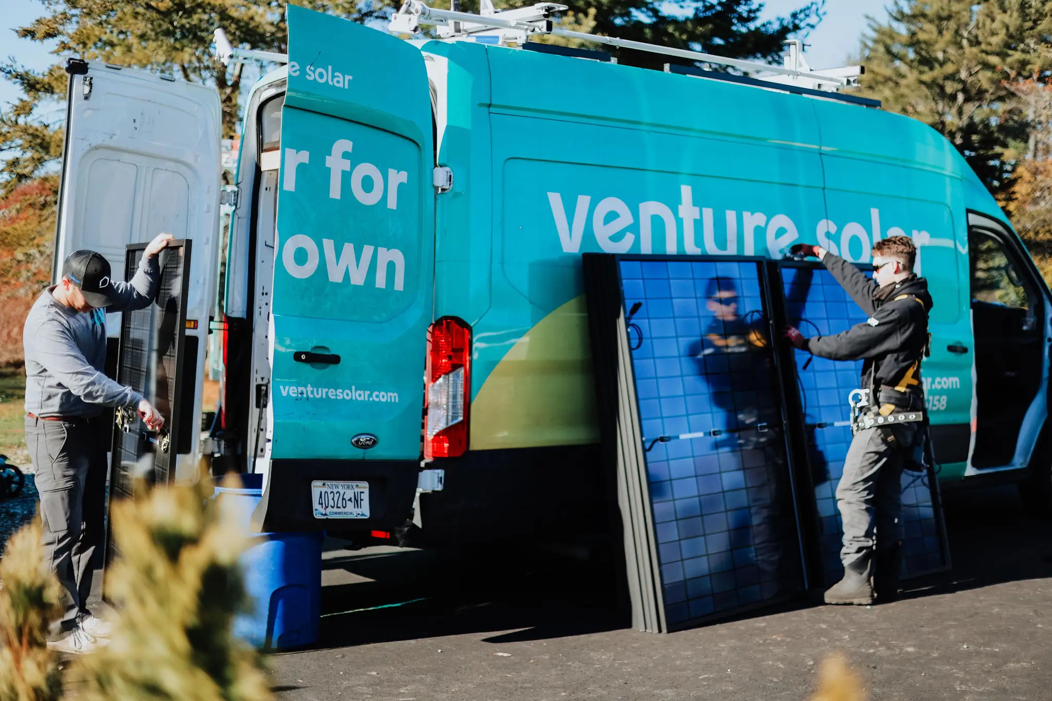 Two workers prepare to install solar panels beside a green Venture Solar van on a sunny day, with trees in the background.