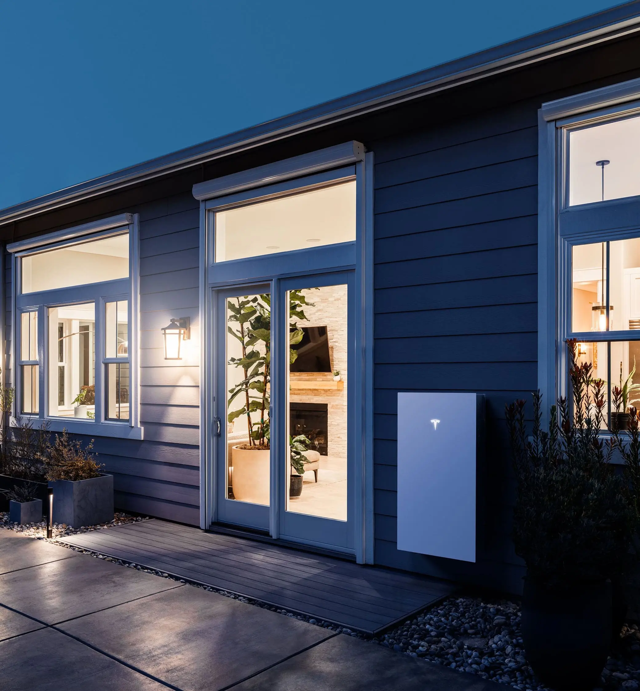 A modern house exterior at dusk with large windows, a lit interior, plants inside, and a white Tesla Powerwall battery mounted on the wall outside.