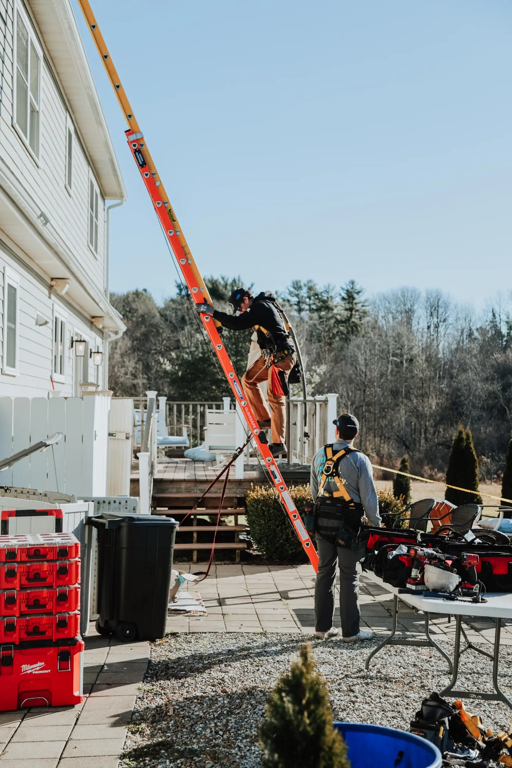 Two workers in safety gear install a Whole Home Solar system outside a house; one stands on an orange ladder, while the other stands by tools and equipment on a table.