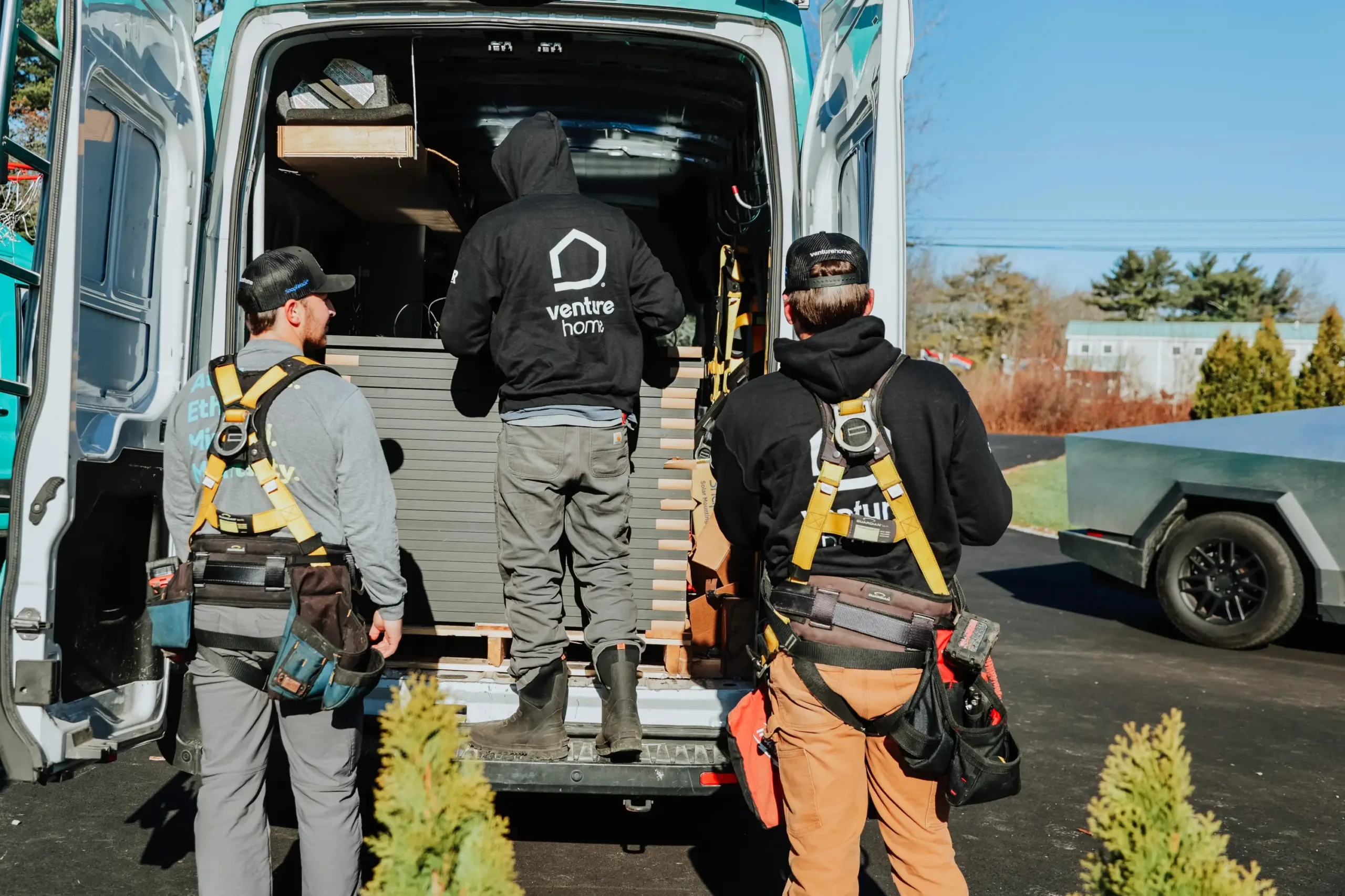 Three workers in safety gear, expert solar installers, stand at the back of a van, loading or unloading building materials on a sunny day.