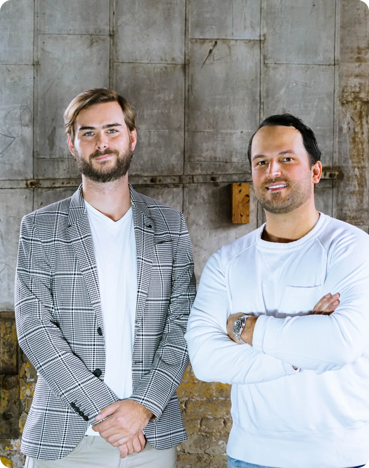 Two men stand side by side in front of a textured, industrial wall, their style as sharp as solar innovation—one in a plaid blazer, the other in a white long-sleeve shirt with arms crossed.
