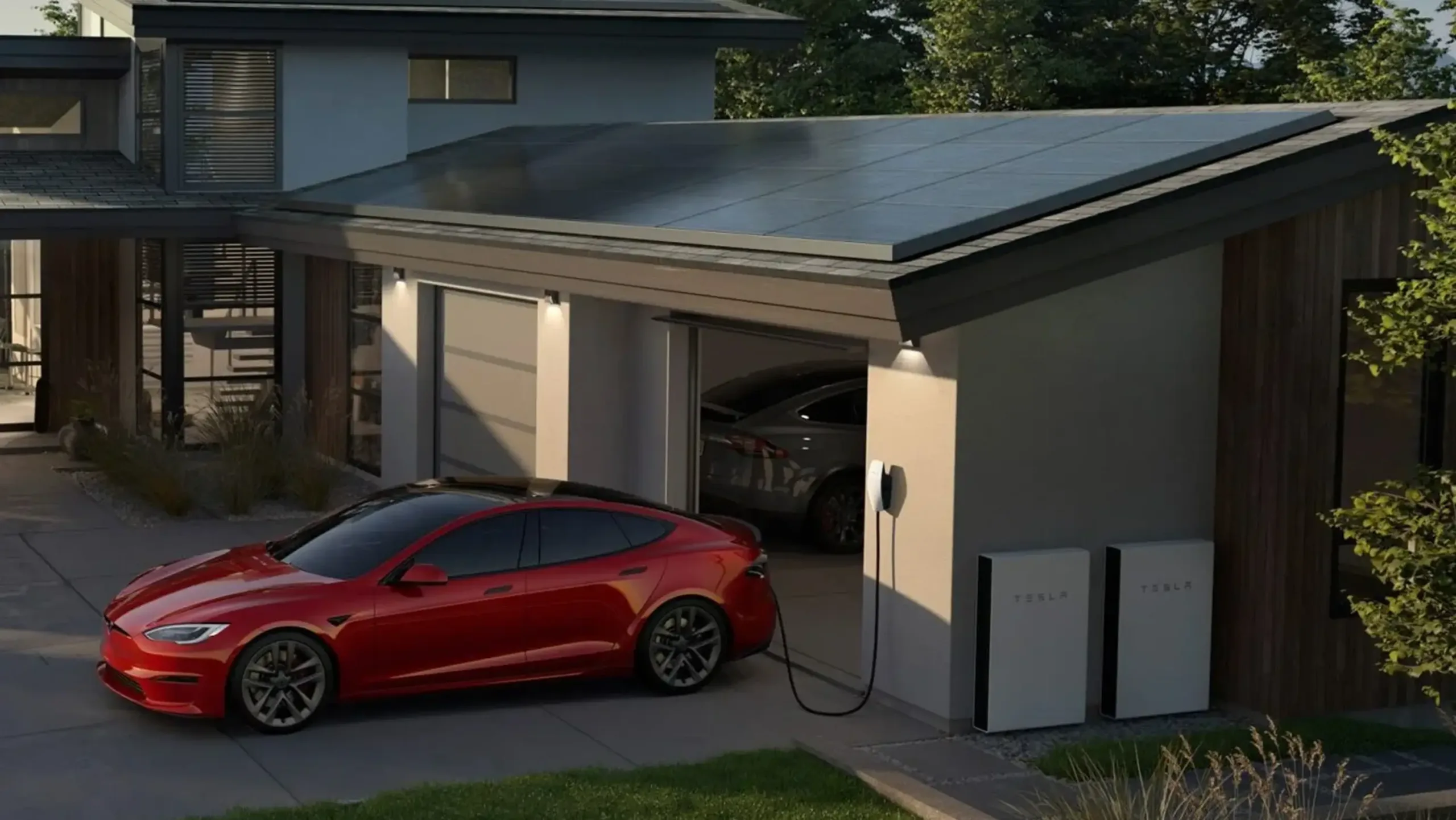 A red electric car is parked in a driveway, charging at a home with solar panels and battery backups, including Tesla Powerwall batteries mounted on the wall.