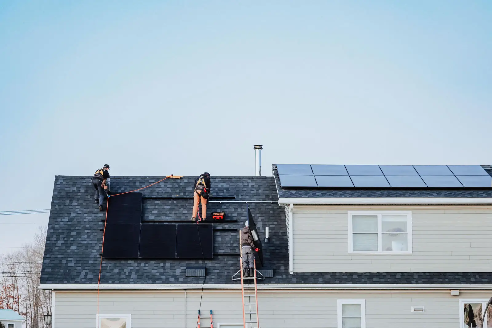 Three workers install solar panels on the roof of a two-story house, as additional solar panels are already mounted on the adjacent section.