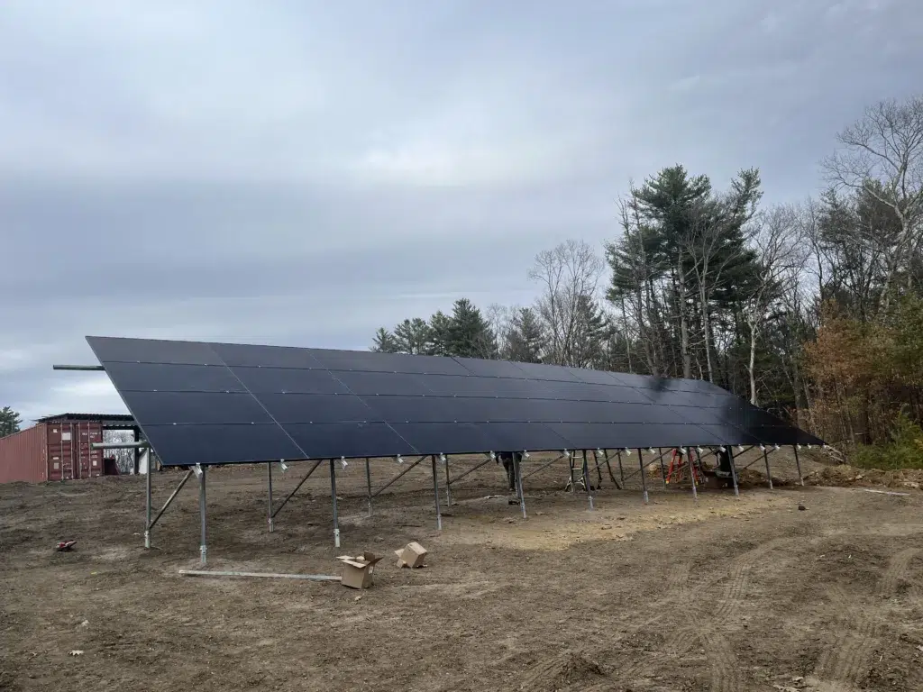 A large solar panel array is installed on a cleared patch of land with trees and overcast sky in the background.
