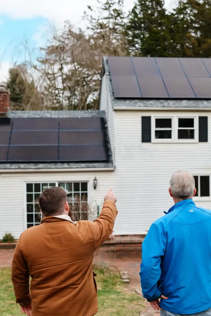 Two people stand outside a white house with solar panels, discussing Massachusetts solar programs. One points toward the panels; trees are visible in the background.